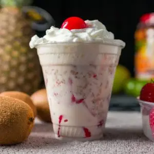 Menú Close-up of a creamy fruit smoothie topped with whipped cream and cherry, surrounded by fresh fruit.